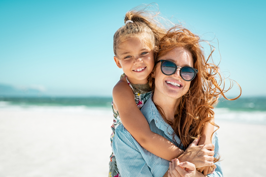 Tochter umschlingt von hinten ihre Arme und Mutter, beide sitzen am Strand